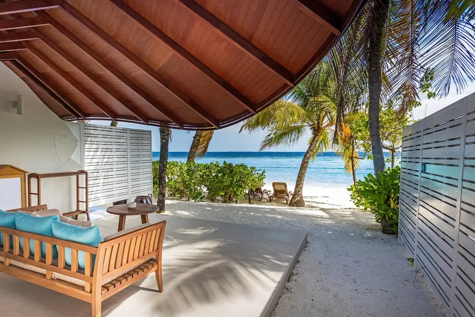 Overdækket terrasse med træsofa og turkise puder ved stranden og palmerne på Machchafushi.