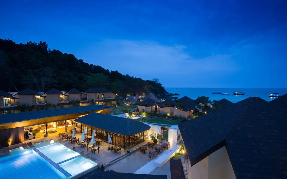 Oversigt over oplyst pool, restaurantterrasse og villaer ved havet i skumringen.