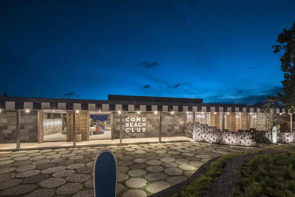Indgangen til COMO Beach Club med skilt og belyst facade ved aftenstid.