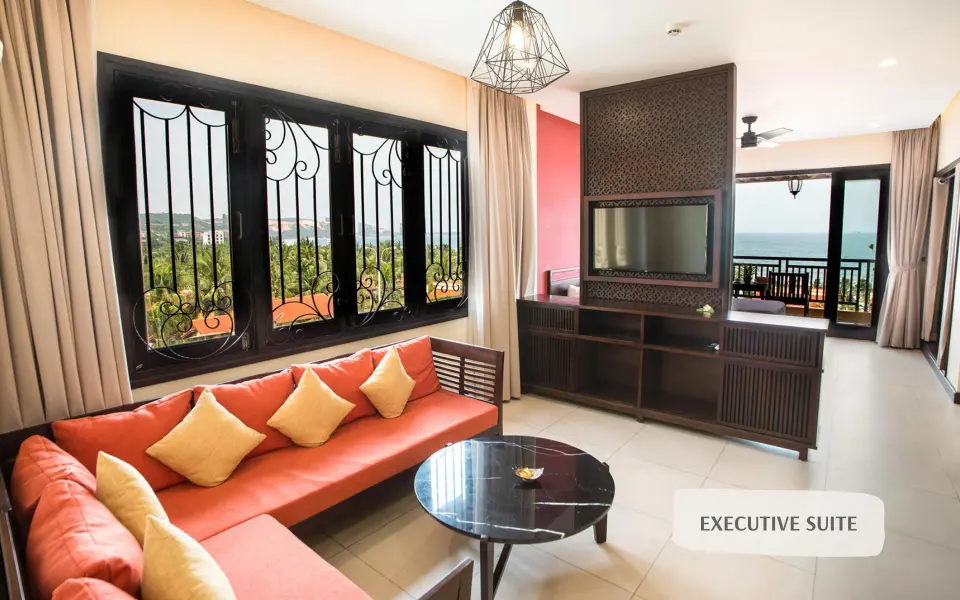 Opholdsstue med orange hjørnesofa, rundt bord og udsigt mod havet.