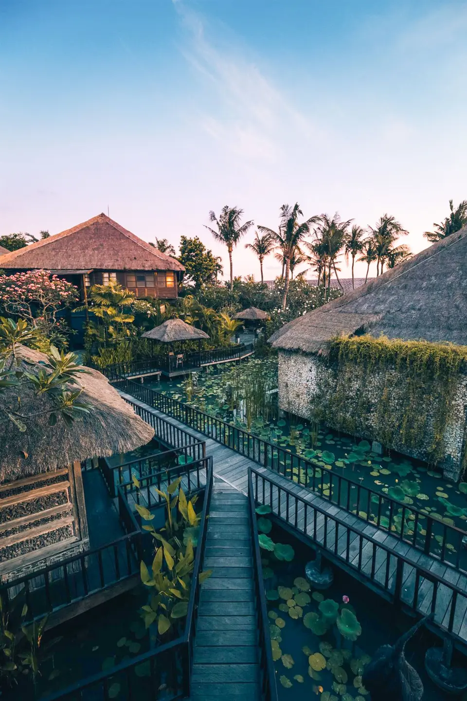 Trægangbro mellem stråtækte bygninger og dam på Hotel Tugu Bali
