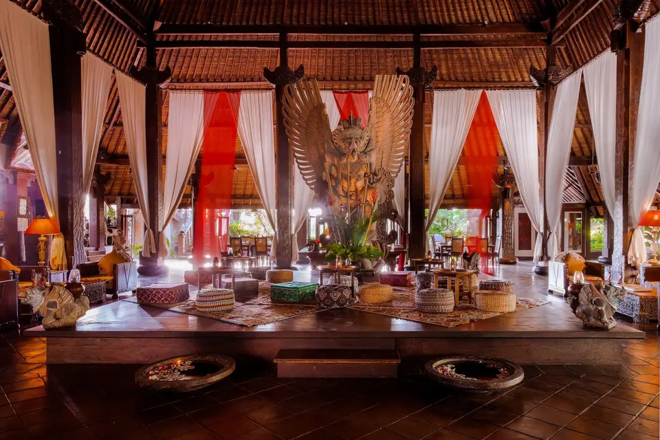 Højloftet fællesrum på Hotel Tugu Bali med træskulptur, siddepuder og lange gardiner.
