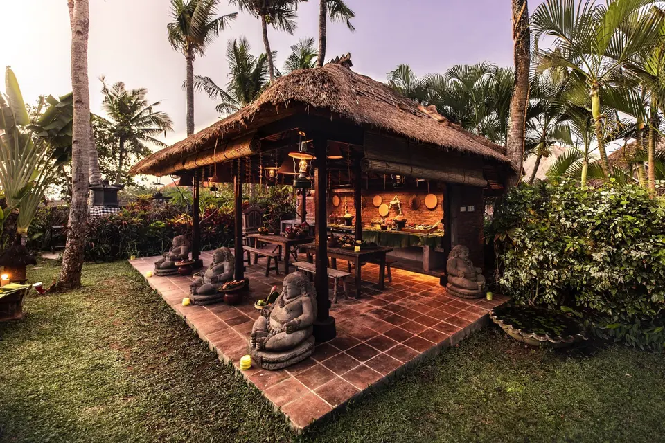 Åben havepavillon på Hotel Tugu Bali med stråtag, træmøbler og stenskulpturer.