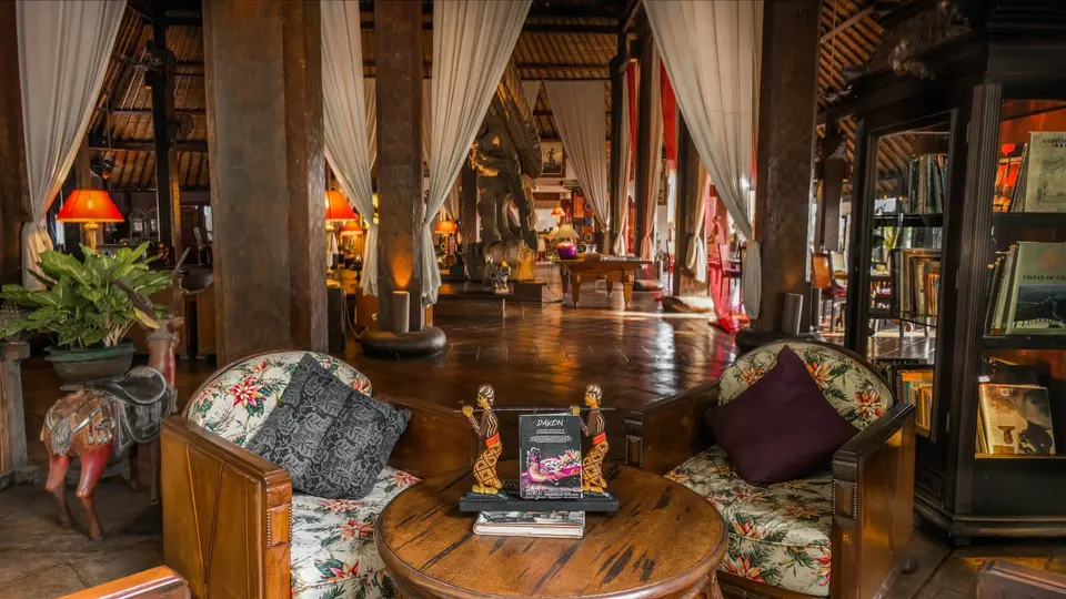 Opholdsrum på Hotel Tugu Bali med blomstrede lænestole, rundt træbord og traditionelle skulpturer.