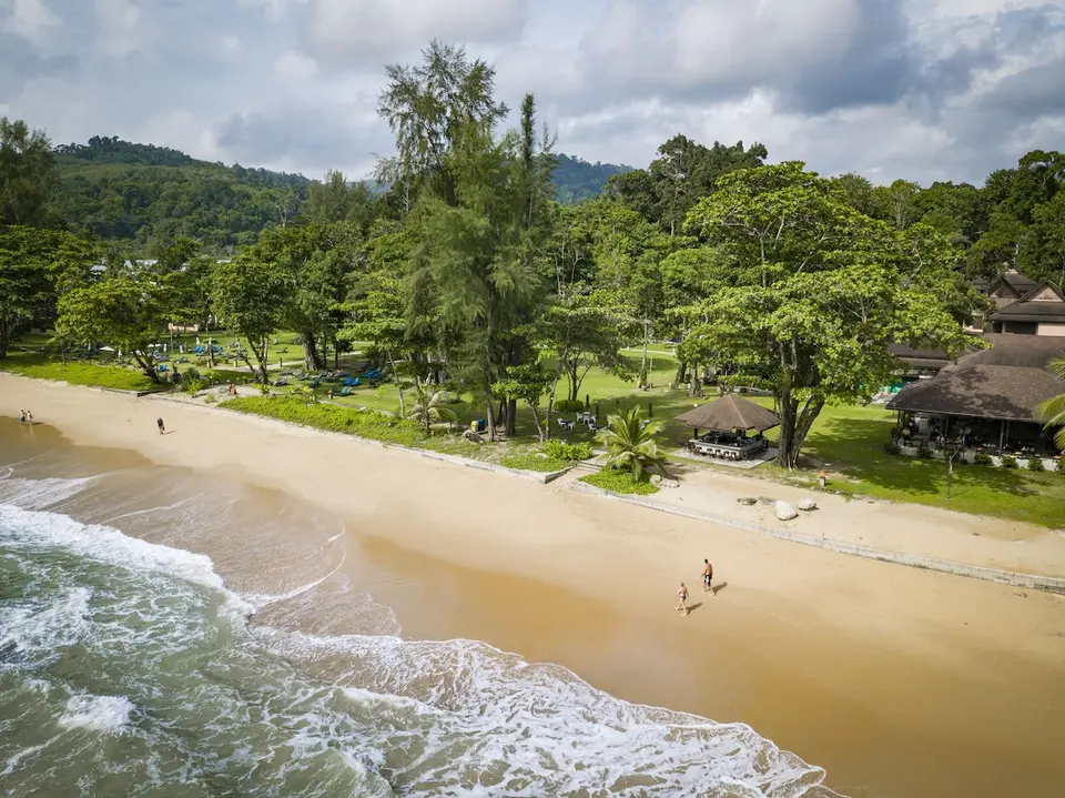 Sandstrand foran Khaolak Merlin Resort med bølger, store træer og resortets grønne have