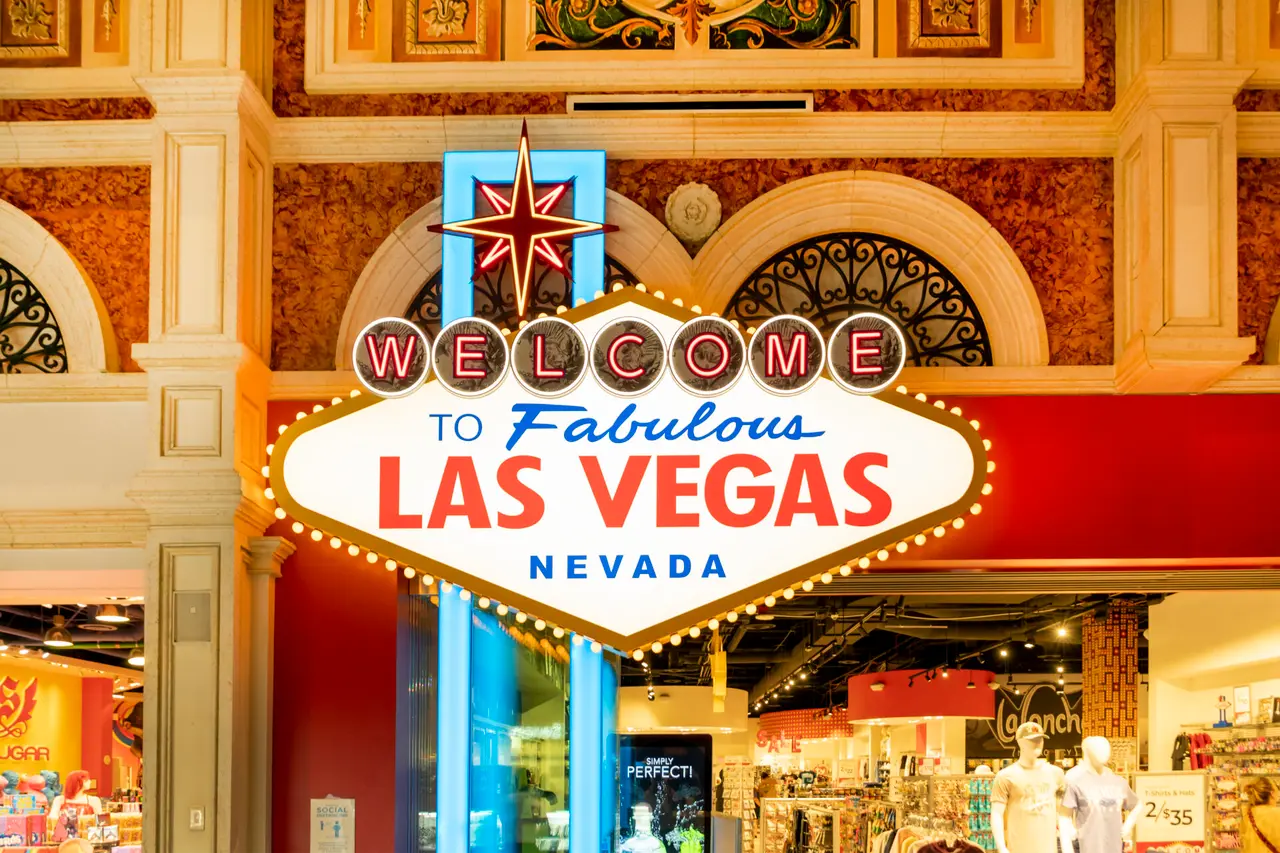 Et oplyst velkomstskilt med teksten Las Vegas foran butikker i en overdækket bygning.