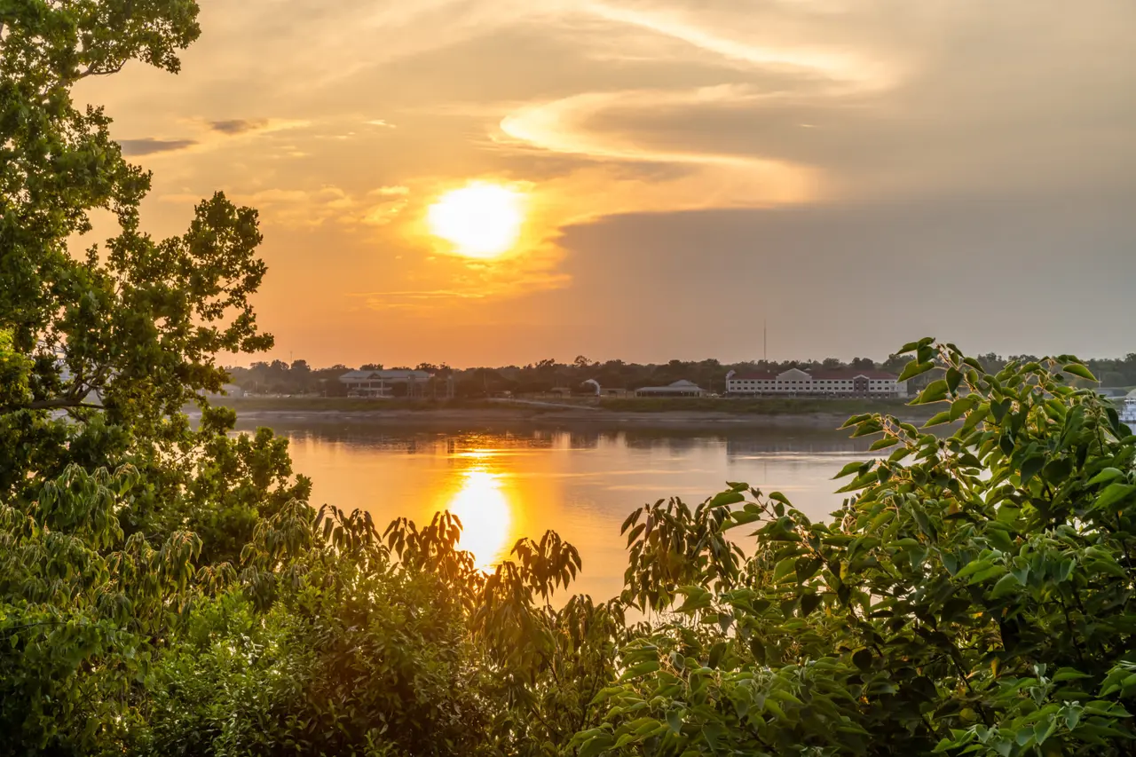 Solen spejler sig i en bred flod med træer i forgrunden og bygninger på den modsatte bred.