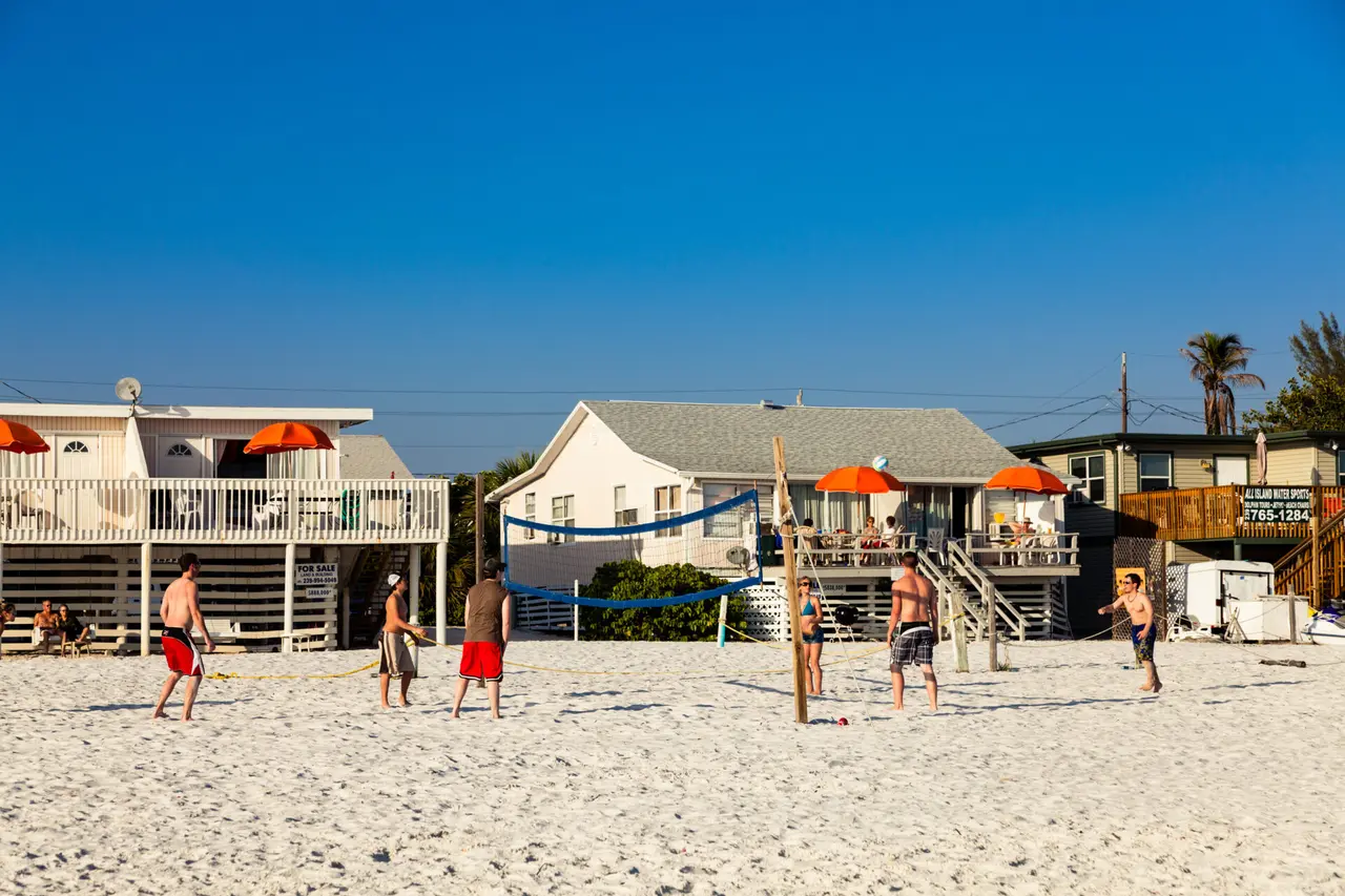 Mennesker spiller beachvolley foran lave strandhuse med terrasser og orange parasoller.