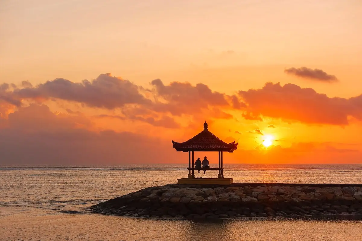 To personer sidder i en pavillon på en stenmole med havet og en lav sol i baggrunden.