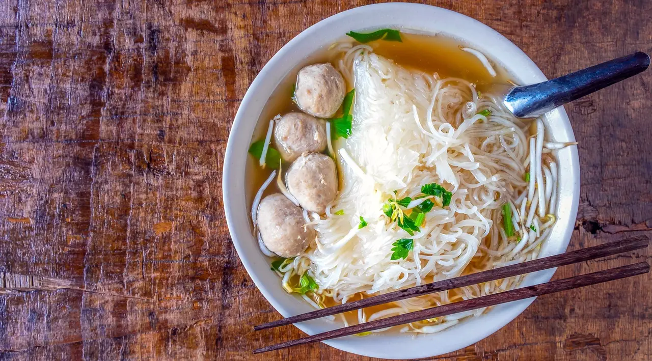 En skål nudelsuppe med boller, bønnespirer og grønt, serveret med ske og spisepinde.