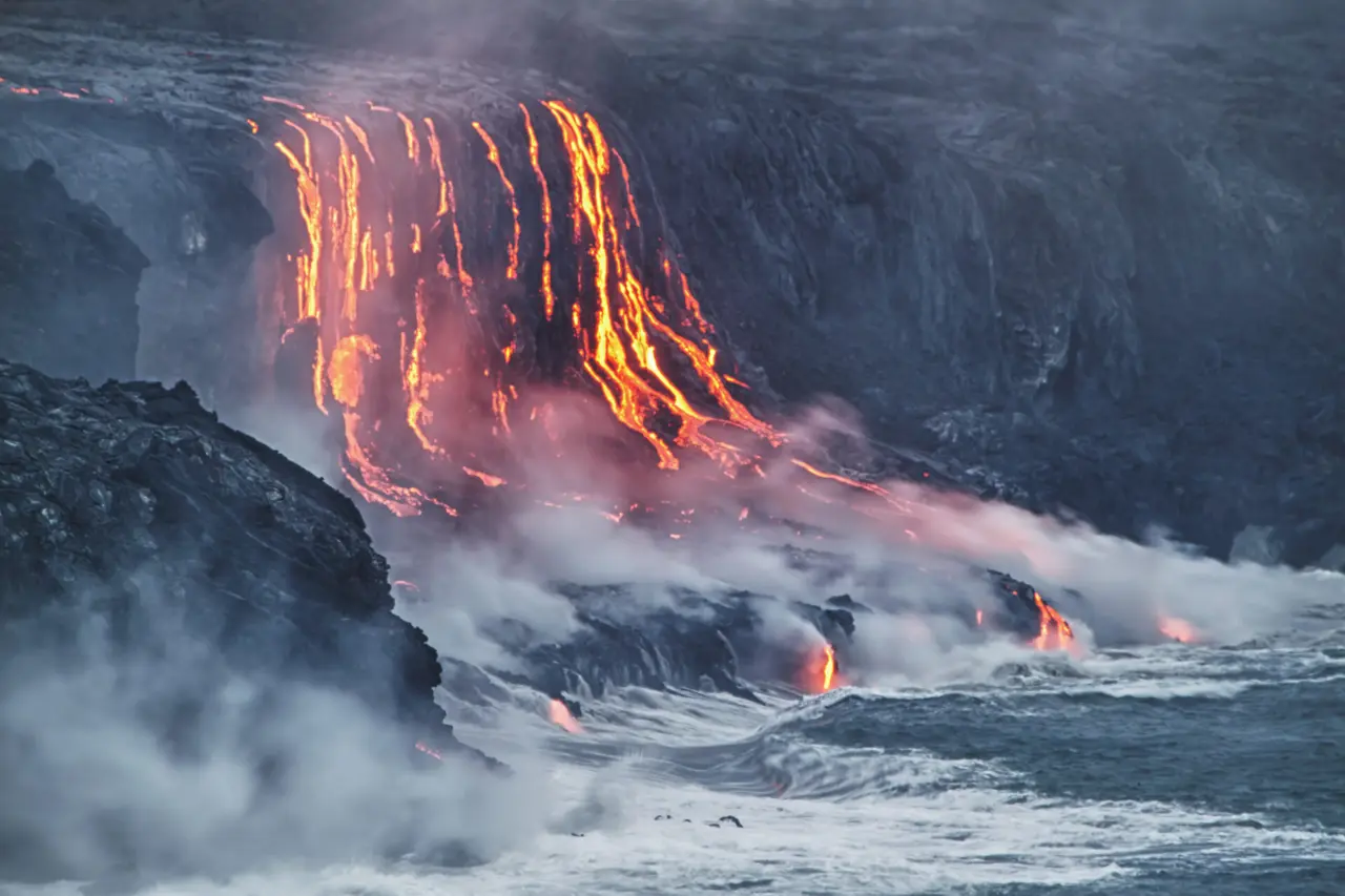 Glødende lava løber over mørke klipper mod havet, hvor damp stiger op.