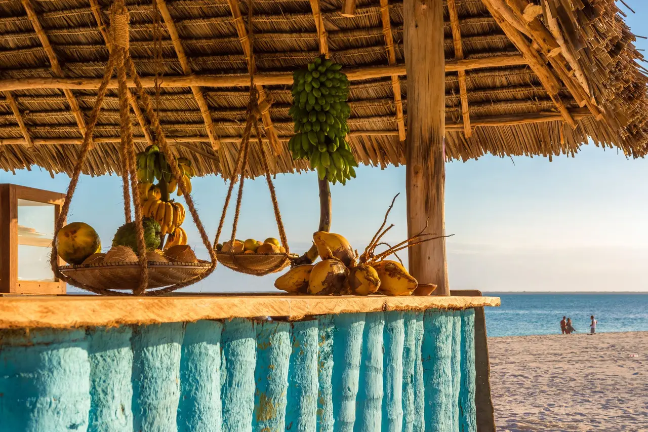 Frugt og kokosnødder ved en turkis bar under stråtag med stranden og havet i baggrunden.