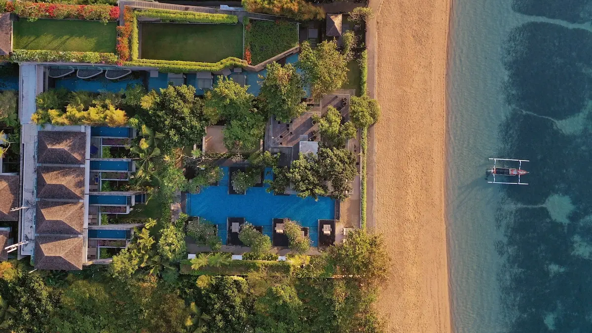 Maya Sanurs bygninger, grønne tage og pools set ovenfra ved sandstranden.