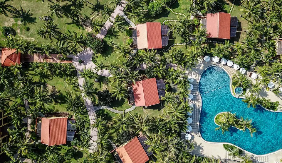 Pool og bungalows med røde tage set ovenfra mellem palmer og havestier.