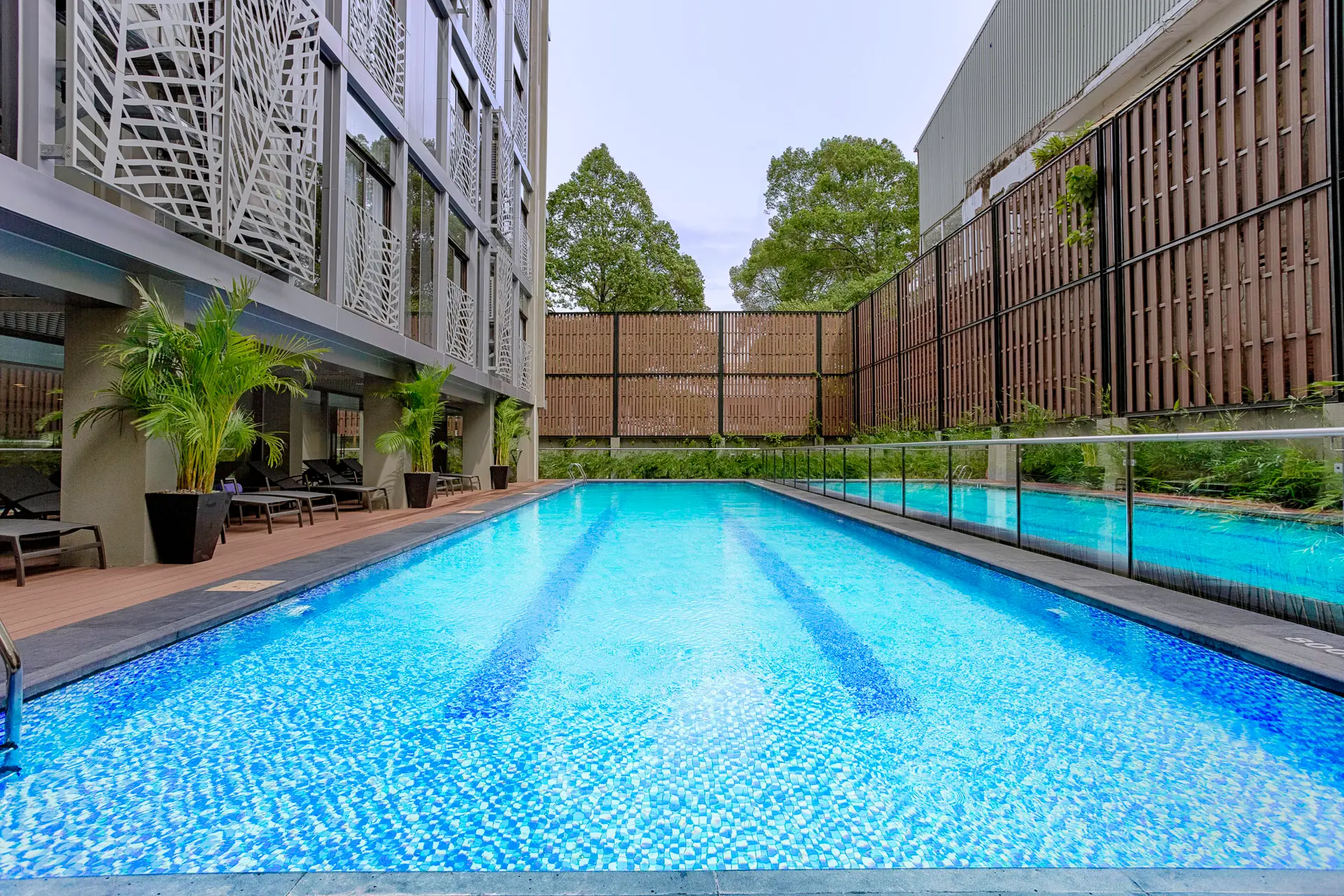 Lang udendørs pool med blå mosaikker, liggestole og hotellets facade.