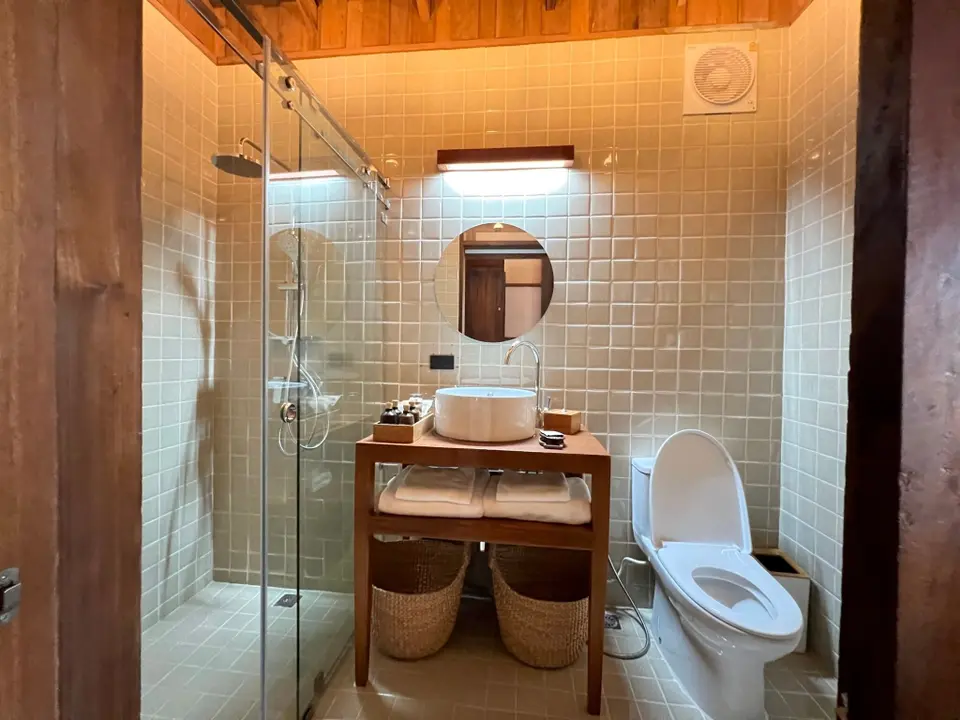 Badeværelse med lyse fliser, glasafskærmet bruser, rund håndvask og toilet