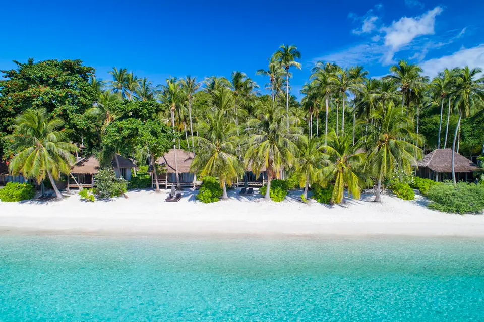 Bungalows og palmer langs en hvid sandstrand set fra havet.