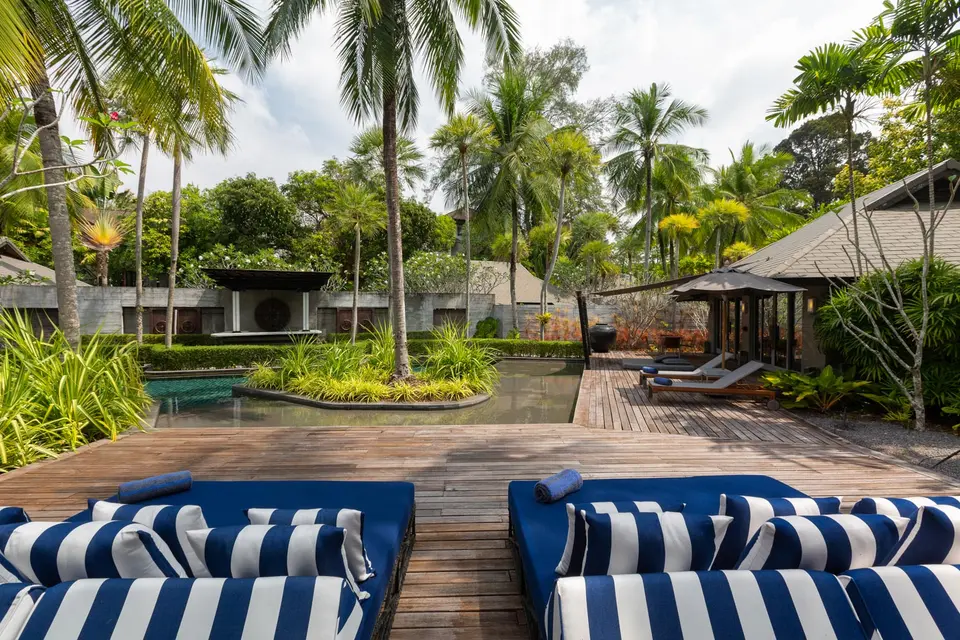 Solrig poolterrasse med trædæk, stribede solsenge og frodige palmer ved moderne hotel på Bali.