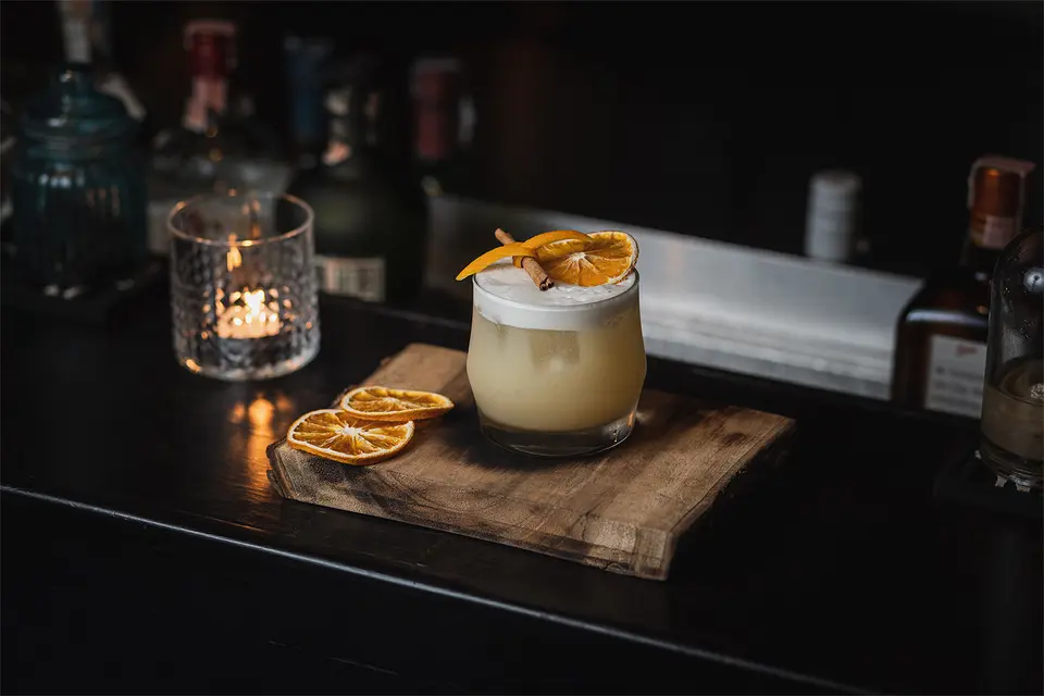 Cocktail med skum og tørret appelsin på et træbræt ved en bar.