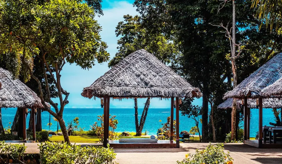 Stråtækt pavillon omgivet af grønne træer ved Khaolak Merlin Resort med udsigt til havet under blå himmel.