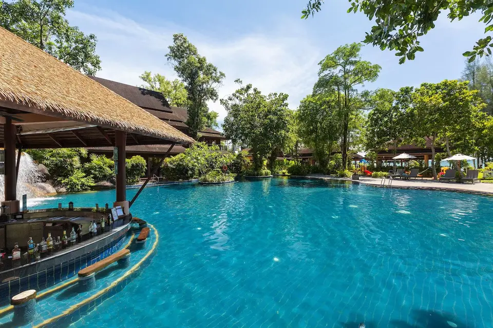 Stor udendørs pool med stråtækt swim-up bar omgivet af tropiske træer på Khaolak Merlin Resort under blå himmel.