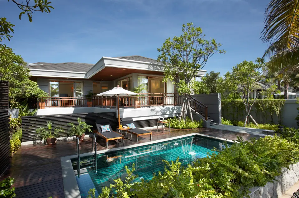 Villa med overdækket terrasse, liggestole og en mindre pool omgivet af grønne planter.