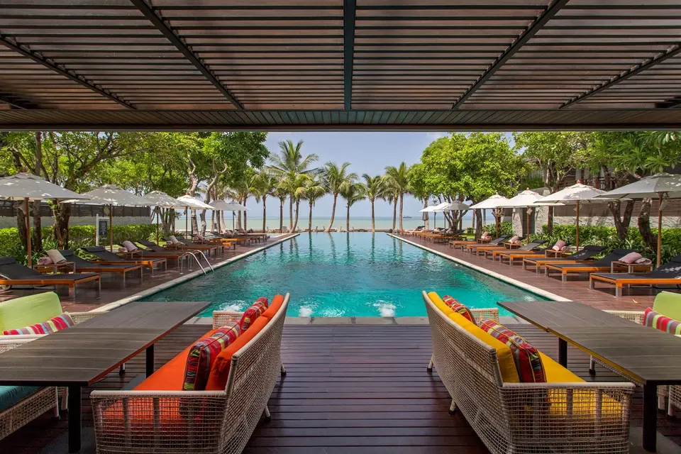 Udendørs pool med liggestole, parasoller og palmer på Rest Detail Hotel Hua Hin set fra skyggefuld terrasse en solrig dag.