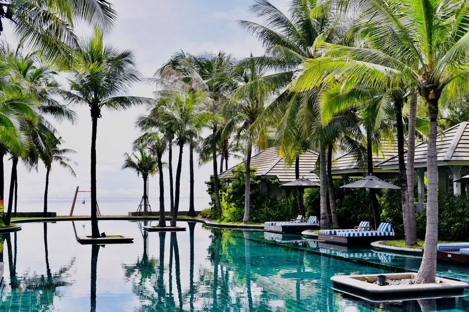 Poolområde med palmer, liggestole og cabanaer ved stranden på Rest Detail Hotel Hua Hin under let skyet himmel.