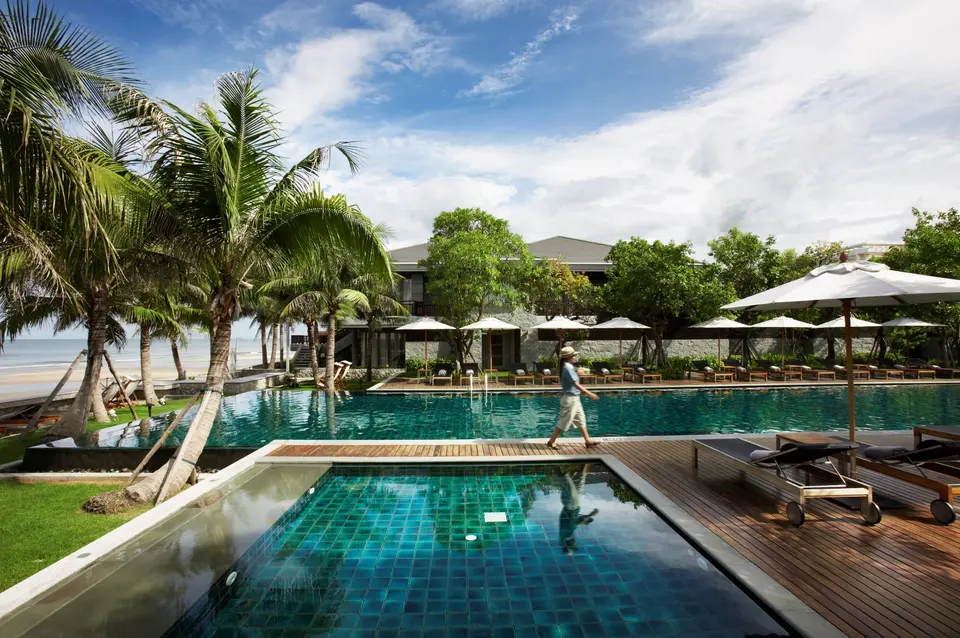 Person spadserer langs moderne pool med palmer og liggestole på Rest Detail Hotel Hua Hin med strand og havudsigt i baggrunden.