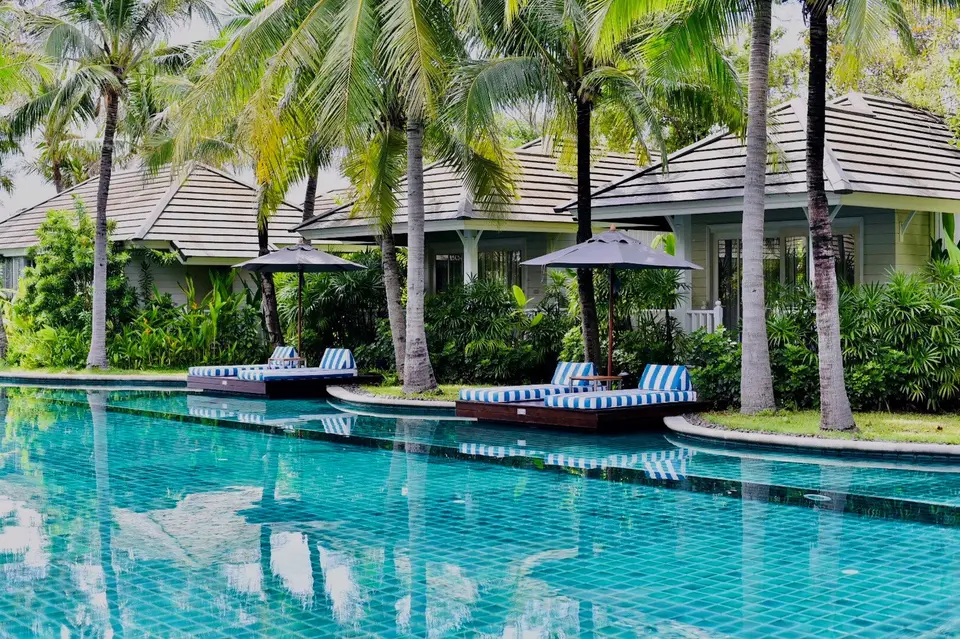 Poolområde med stribede liggestole og parasoller ved Rest Detail Hotel i Hua Hin, omgivet af palmer og tropisk have.