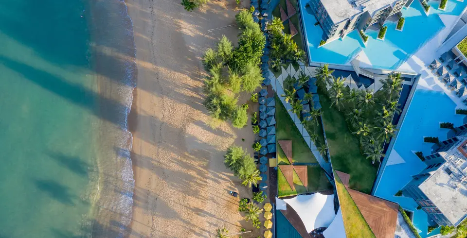 Sandy strand med turkisblåt hav og La Vela Khao Laks moderne resort omgivet af grønne træer og elegante pools.