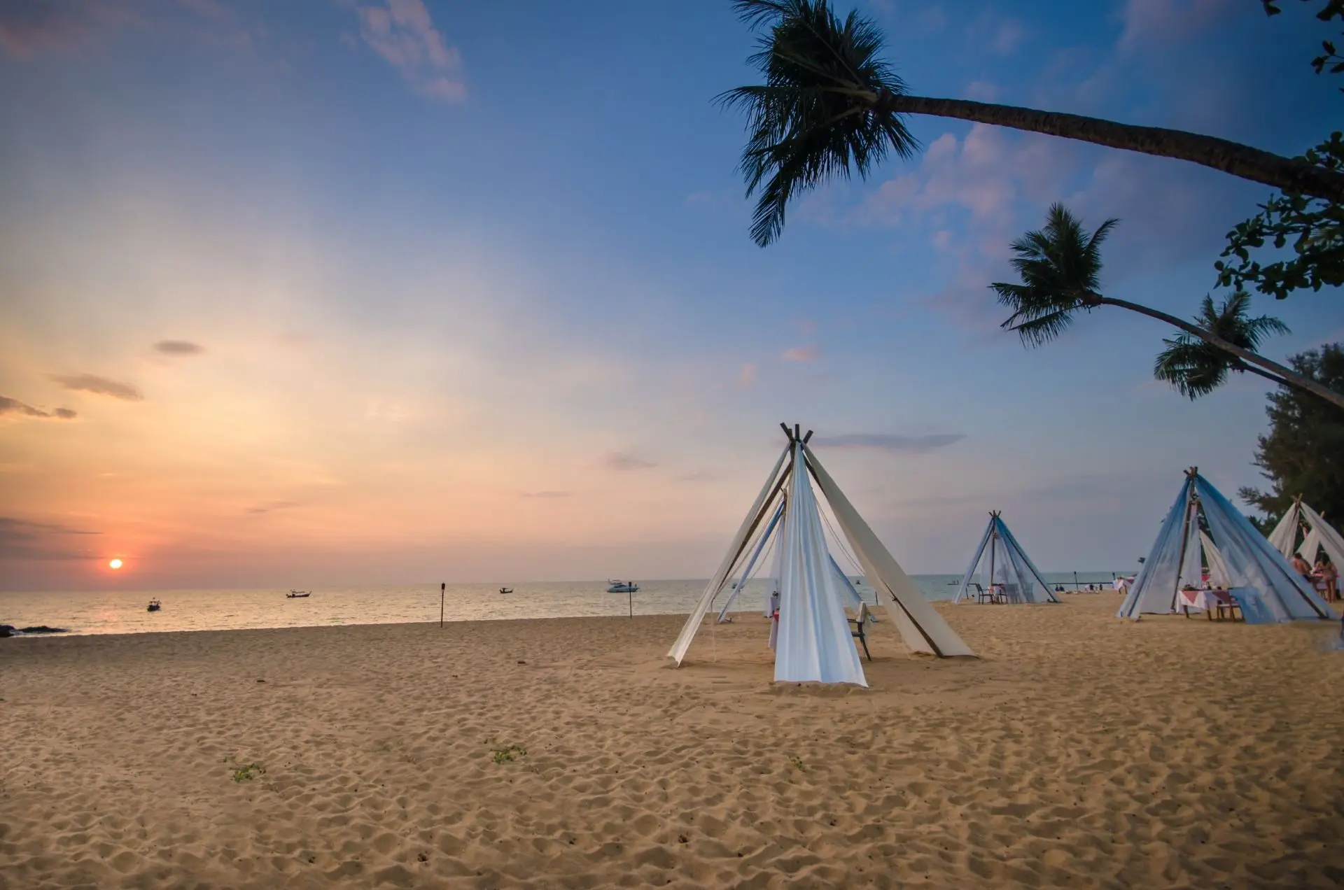 Hvide telte på sandstrand ved solnedgang nær La Vela Khao Lak, med palmer og både på det rolige have i baggrunden.