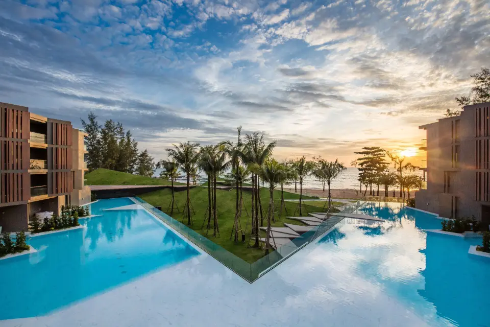 Moderne resort og stor pool omgivet af palmer ved stranden i Khao Lak, badet i varm solnedgangsstemning.