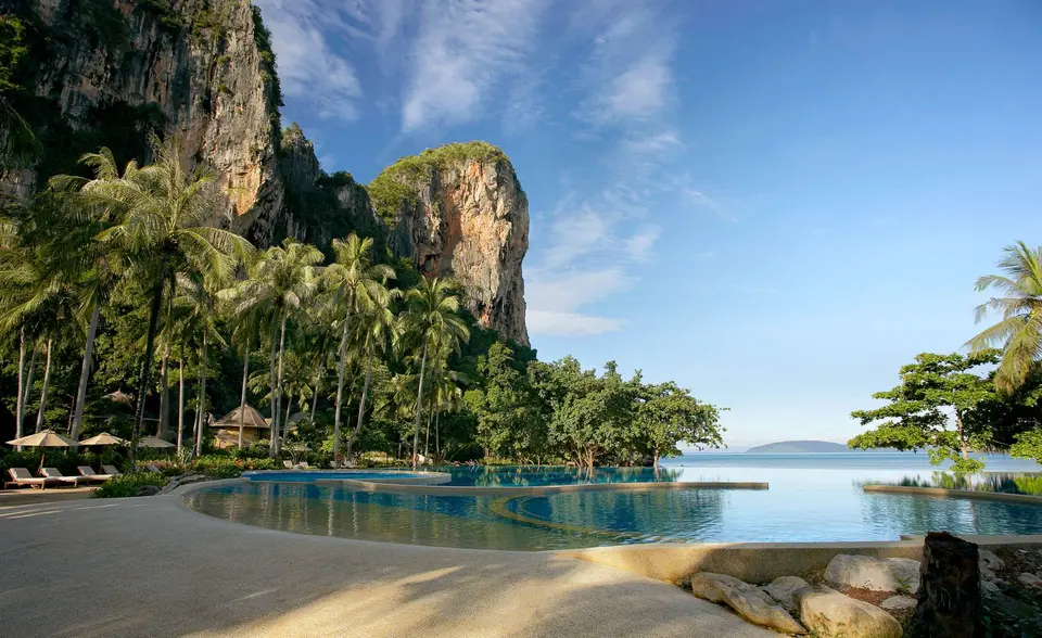 Luksuspool med bløde kurver omgivet af palmer og klipper, med udsigt til havet under blå himmel i Rayavadee, Krabi.
