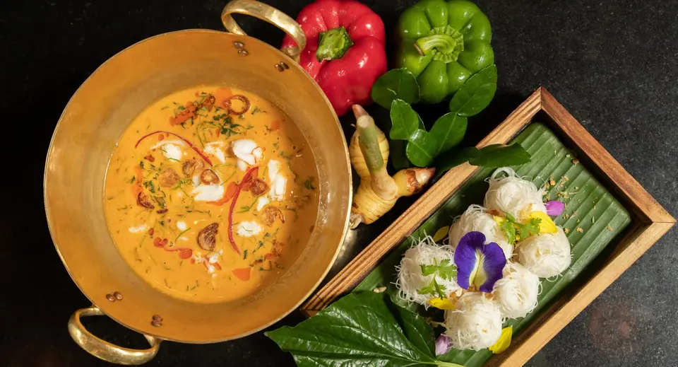 Orange sauce med urter i en metalskål og små portioner nudler pyntet med blomster.