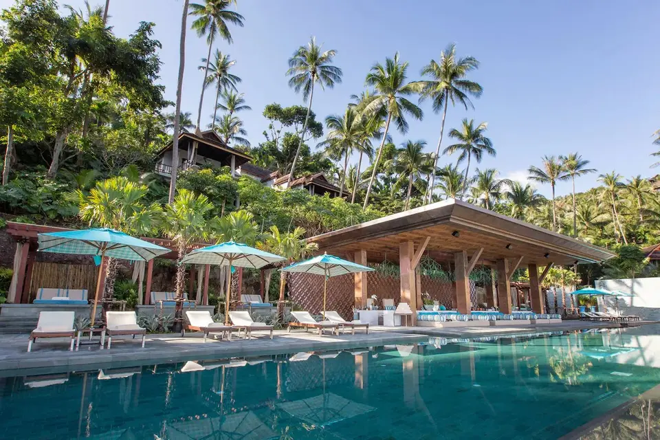 Luksuspool med hvide liggestole og turkise parasoller omgivet af palmer på Four Seasons Resort Koh Samui.