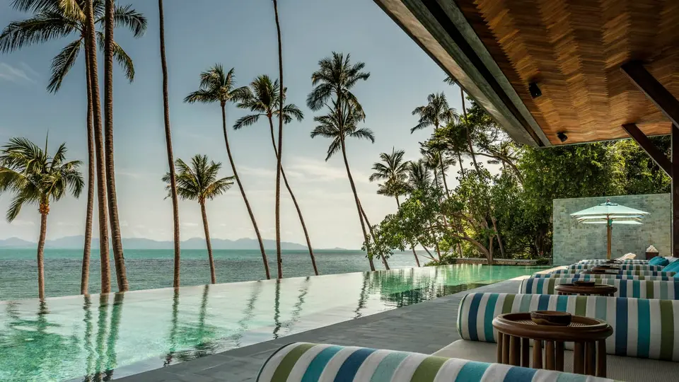 Infinity pool med stribede liggestole og havudsigt på Koh Samui resort, omgivet af palmer og afslappet feriestemning.