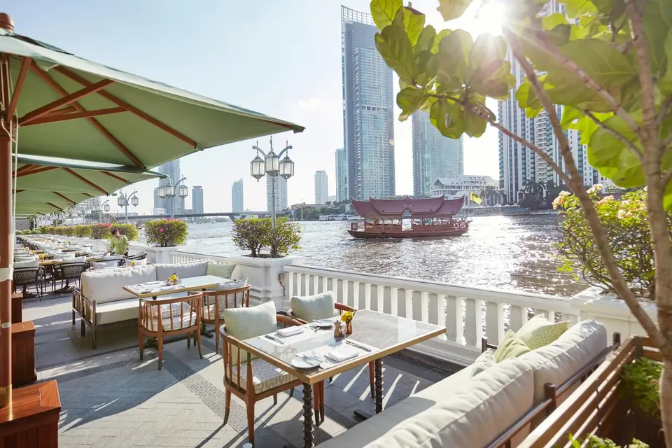 Udendørs restaurant ved floden på Mandarin Oriental Bangkok med udsigt til båd, grønne træer og byens skyline i solskin.