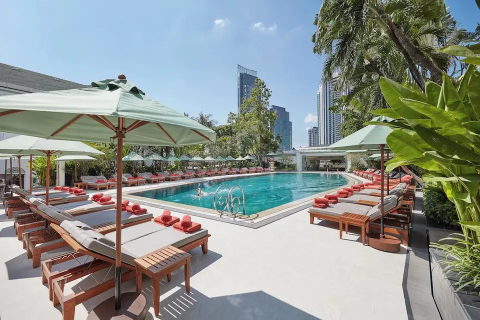 Afslappende poolområde på Mandarin Oriental Bangkok med grønne parasoller, røde håndklæder og frodig have midt i byen.