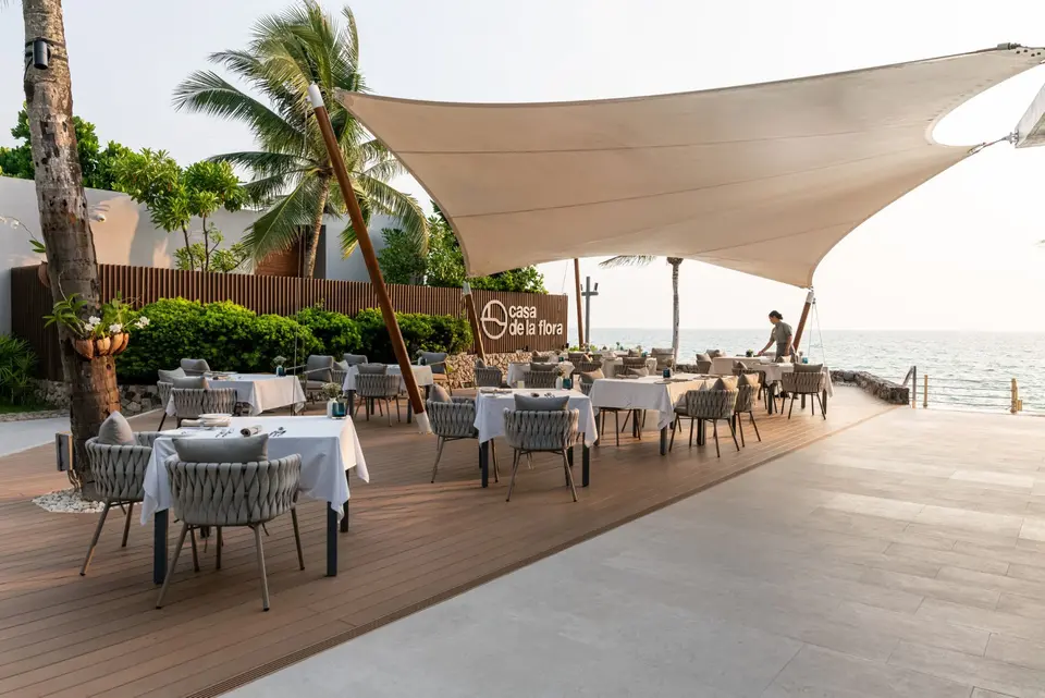 Udendørs restaurant ved havet på Casa de la Flora, hvor tjener dækker borde under hvide sejl blandt palmer og frodig natur.