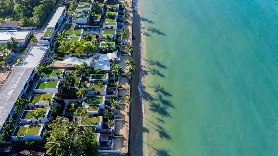 Strandvillaer med grønne tage og palmer langs sandstrand ved det turkisblå hav i Khao Lak, Thailand.