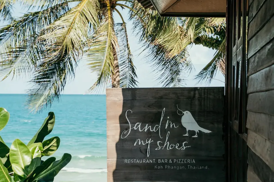Træskilt foran strandbar omgivet af palmer og bølger på Koh Phangan skaber en afslappet feriestemning.