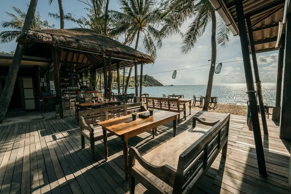 Træmøbleret restaurant under palmer ved strandkanten på Beach Loft, med udsigt over havet i solskin.