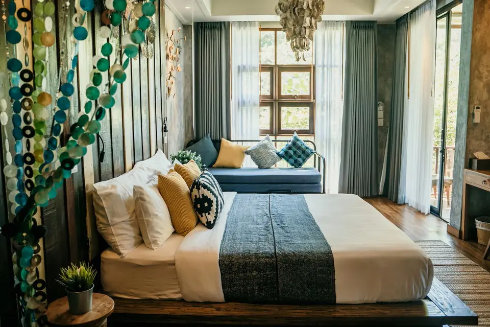 Hyggeligt beach-loft soveværelse med bløde puder, sofa ved store vinduer og sollys gennem lette gardiner.